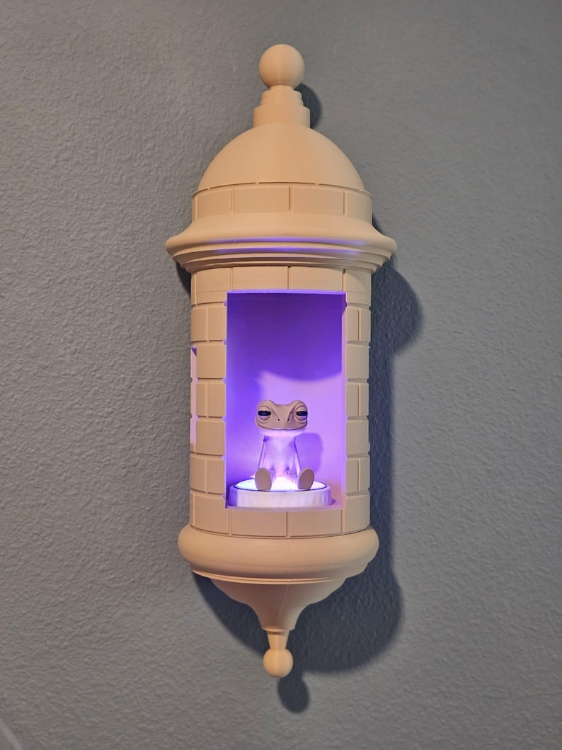 May include: A beige, castle-shaped wall sconce with a small frog figurine inside, illuminated by a purple light. The sconce has a domed top and a decorative base, mounted on a blue wall.