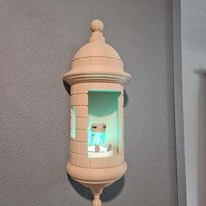 May include: A beige-colored, decorative wall sconce shaped like a small tower. Inside, a small frog figurine sits on a platform, illuminated by a soft green light. The sconce is mounted on a gray wall.