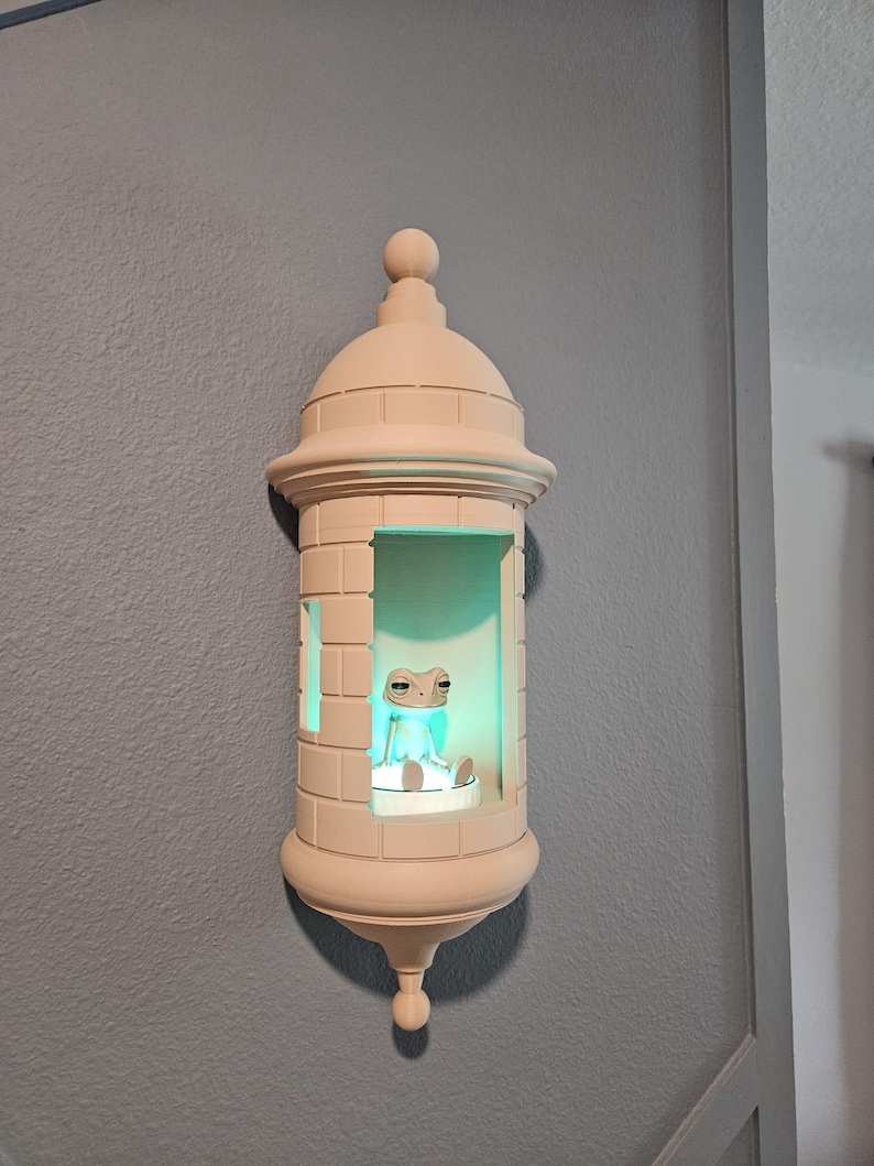 May include: A beige-colored, decorative wall sconce shaped like a small tower. Inside, a small frog figurine sits on a platform, illuminated by a soft green light. The sconce is mounted on a gray wall.