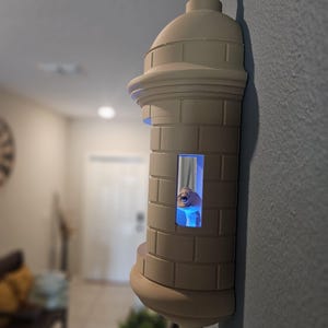 May include: A beige, 3D-printed wall sconce shaped like a fortress tower. The tower has a domed top, a cylindrical body with rectangular details, and a small window with a blue light. The sconce is mounted on a gray wall.
