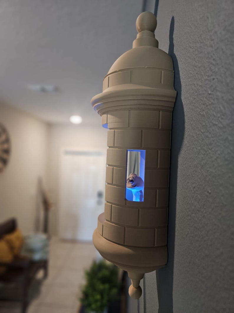 May include: A beige, 3D-printed wall sconce shaped like a fortress tower. The tower has a domed top, a cylindrical body with rectangular details, and a small window with a blue light. The sconce is mounted on a gray wall.