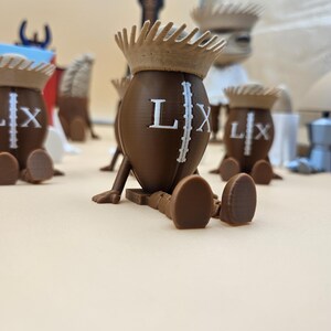 May include: Brown 3D-printed figures shaped like footballs, each with a crown and the letters "LX". The figures have legs and feet, and are arranged on a light-colored surface. Other objects are in the background.