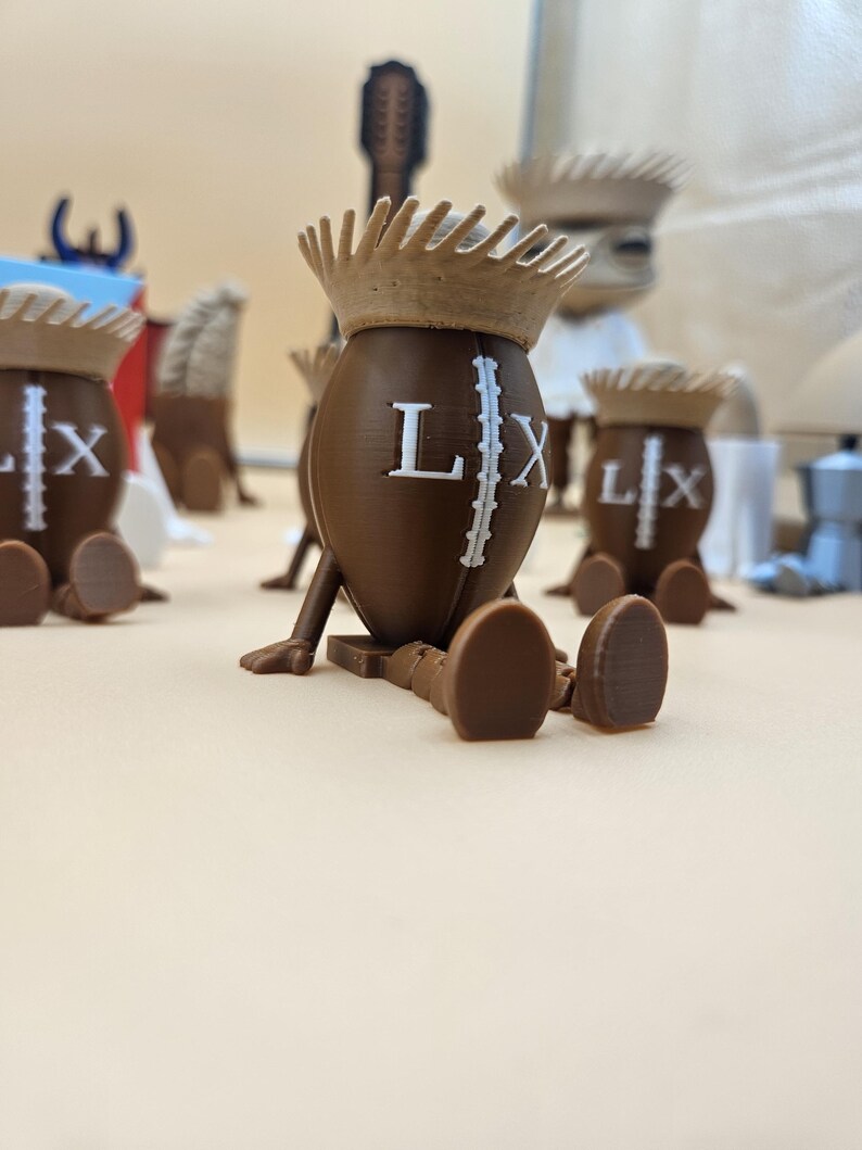 May include: Brown 3D-printed figures shaped like footballs, each with a crown and the letters "LX". The figures have legs and feet, and are arranged on a light-colored surface. Other objects are in the background.