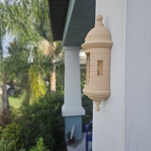 May include: A beige, decorative wall-mounted mailbox shaped like a miniature fort. The mailbox has a domed top, brick-like detailing, and a small opening for mail. It is mounted on a white exterior wall.