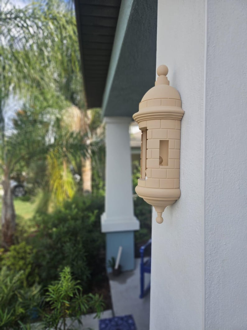 May include: A beige, decorative wall-mounted mailbox shaped like a miniature fort. The mailbox has a domed top, brick-like detailing, and a small opening for mail. It is mounted on a white exterior wall.