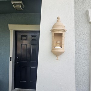 May include: A beige, decorative wall-mounted lantern with a domed top and a small, open compartment. The lantern is mounted on a textured white wall next to a black door. A small statue is inside the lantern.