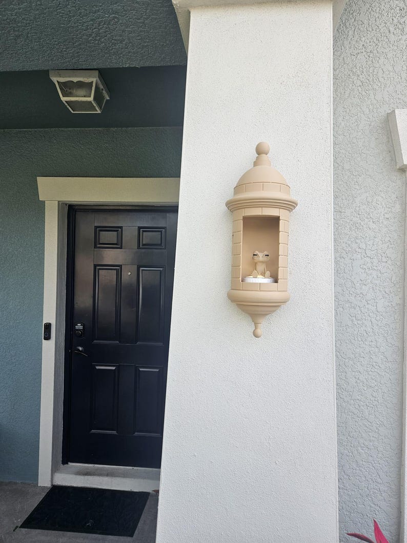 May include: A beige, decorative wall-mounted lantern with a domed top and a small, open compartment. The lantern is mounted on a textured white wall next to a black door. A small statue is inside the lantern.