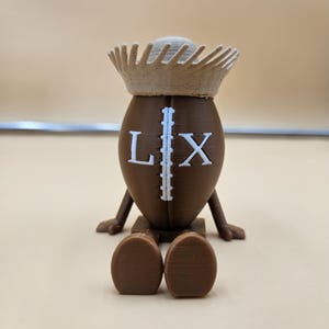 May include: A brown, football-shaped figurine with the letters "L" and "X" on the front. The figurine has a crown and small legs and feet. The figurine is sitting on a brown base.