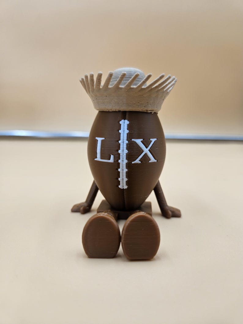 May include: A brown, football-shaped figurine with the letters "L" and "X" on the front. The figurine has a crown and small legs and feet. The figurine is sitting on a brown base.