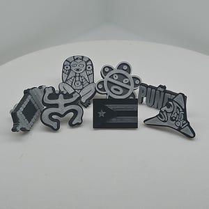 May include: A collection of black and gray 3D-printed charms featuring various designs. Included are a stylized figure, a flower-like shape, a Puerto Rican flag, and a charm with the word "PUN".