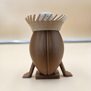 May include: A brown, egg-shaped figurine with a crown. The figurine has small legs and feet, and is standing on a square base. The crown is a light beige color and has a spiky design.