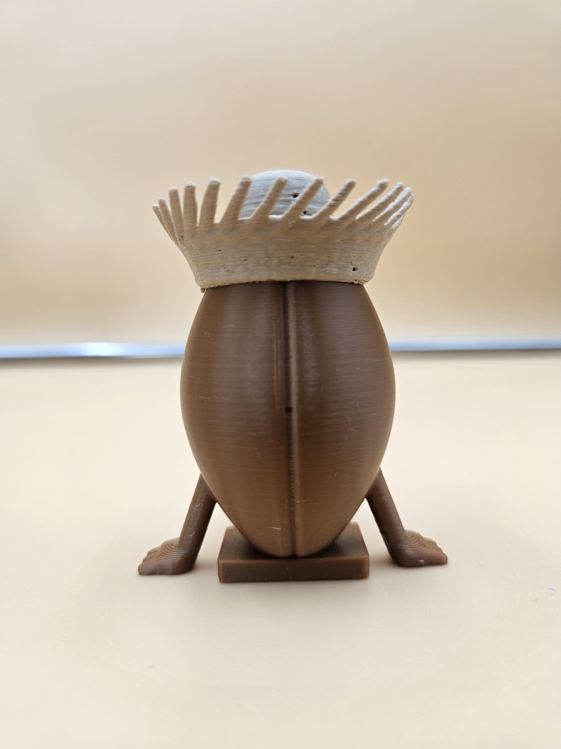May include: A brown, egg-shaped figurine with a crown. The figurine has small legs and feet, and is standing on a square base. The crown is a light beige color and has a spiky design.