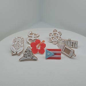 May include: A collection of small, decorative charms featuring various designs. Included are a red hibiscus flower, a Puerto Rican flag, and several tan-colored charms with intricate patterns and the word "PUÑETA".