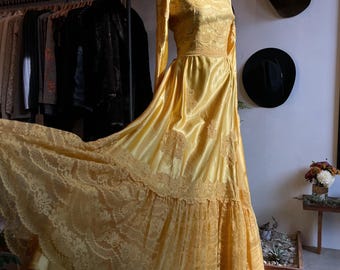 May include: A vintage, floor-length yellow dress with long sleeves. The dress features a lace bodice and a flowing skirt with lace trim. The dress is displayed on a mannequin in a boutique setting, with hats and clothing racks in the background.
