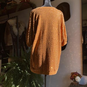 Puede incluir: Vestido naranja quemado con lentejuelas y mangas cortas, exhibido en un maniquí. El vestido está cubierto de pequeñas lentejuelas redondas que captan la luz. Tiene un escote redondo y un corte holgado y cómodo.