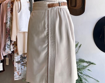 May include: A beige wrap skirt with a brown leather belt and brown stitching detail. The skirt is knee-length and has a straight silhouette.