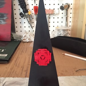 May include: A black triangular pyramid with a red geometric symbol on the front. The symbol is a square with a triangle inside, and the triangle has a circle inside.