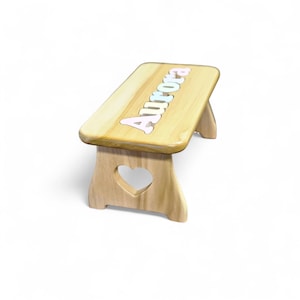 May include: A small wooden bench with a light natural wood grain finish. The top surface features the name Aurora in raised, pastel-colored letters. The side supports are designed with a heart-shaped cutout in the center of each panel.