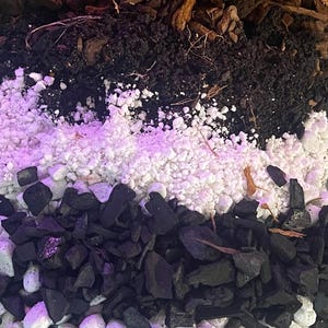 May include: A layered composition of various horticultural substrates. The top layer features coarse, brown bark chips, followed by dark soil, white perlite, black charcoal, and rounded white pebbles. The bottom layer consists of reddish-brown clay balls.