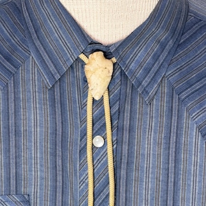 Puede incluir: Camisa azul a rayas con dos bolsillos en el pecho y botones blancos. La camisa presenta una corbata bolo con un cordón color crema y un colgante de punta de flecha de piedra. La camisa se exhibe en un maniquí.