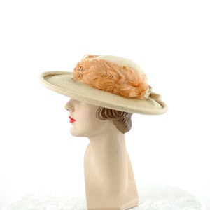 May include: A beige hat with a wide brim, adorned with peach-coloured feathers. The hat is displayed on a mannequin head with red lipstick, showcasing a vintage style.