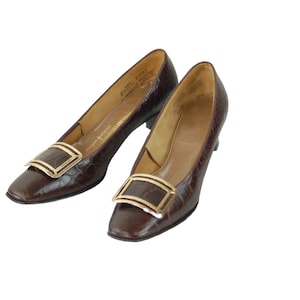 May include: A pair of dark brown pumps with a textured crocodile-embossed finish. Each shoe features a square toe and a prominent, polished gold-tone metal buckle ornament on the vamp. The interior is lined in tan leather with visible branding.