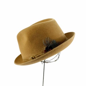 May include: A tan fedora hat with a dark feather accent. The hat has a classic shape with a slightly curved brim and a band around the crown. The feather is black and red. The hat is displayed on a wire stand.