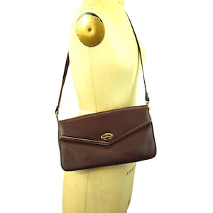 May include: A brown leather shoulder bag with a gold-colored clasp and trim. The bag is rectangular with a flap closure and a long, thin strap. The bag is displayed on a mannequin.