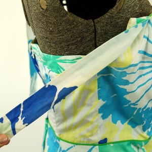 May include: A sleeveless dress featuring a vibrant floral pattern in shades of blue, turquoise, and yellow on a white background. The garment includes a thin green piped trim along the waistline and a matching fabric strap extending from the shoulder.