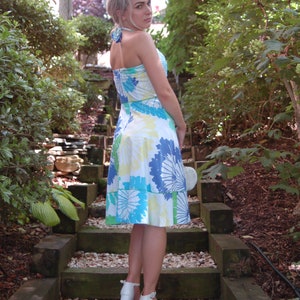 May include: A knee-length halter dress featuring a white base with a vibrant floral pattern in shades of blue, green, and yellow. The dress has a fitted bodice, a flared skirt, and a tie-neck closure. Paired with white block-heeled sandals.