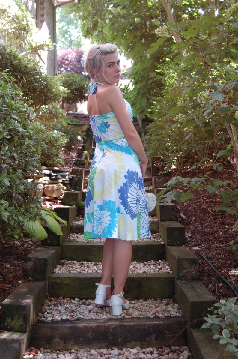 May include: A knee-length halter dress featuring a white base with a vibrant floral pattern in shades of blue, green, and yellow. The dress has a fitted bodice, a flared skirt, and a tie-neck closure. Paired with white block-heeled sandals.