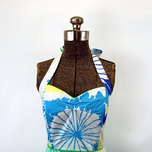 May include: A halter-style dress featuring a white base with a bold, abstract floral print in shades of bright blue, yellow, and green. The bodice has a sweetheart neckline with green piping trim and wide straps that tie behind the neck.
