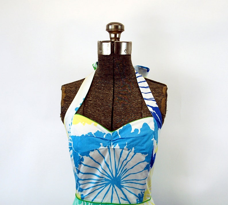 May include: A halter-style dress featuring a white base with a bold, abstract floral print in shades of bright blue, yellow, and green. The bodice has a sweetheart neckline with green piping trim and wide straps that tie behind the neck.