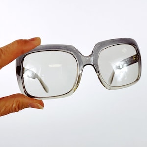 May include: Clear-lensed eyeglasses with a gray, square frame. The glasses are held up against a white background. The lenses are clear, and the frame is a translucent gray color.