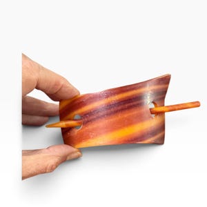May include: A hair slide featuring a rectangular, curved piece with a marbled pattern of orange, brown, and yellow tones. The accessory includes two circular cutouts on the sides, through which a solid, smooth, orange-colored hair stick is inserted.