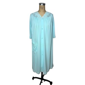 May include: A light blue housecoat featuring a button-down front, three-quarter length sleeves, and a single patch pocket. The garment is accented with delicate white lace trim along the V-neckline, sleeve cuffs, and the bottom hemline.