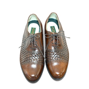 May include: A pair of brown leather oxford shoes featuring a woven texture on the quarter panels, a smooth cap toe, and dark brown laces. The interior features a green label with the text FLORSHEIM. The shoes have a low heel and a rounded toe.