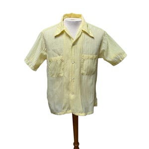 Puede incluir: Una camisa amarilla clara de manga corta con un patrón de rayas verticales texturizadas. La camisa tiene cuello, dos bolsillos delanteros y se abrocha por delante. La camisa se exhibe en un maniquí de madera.