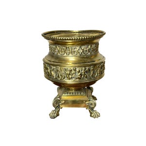 May include: A gold-colored, ornate brass urn with intricate relief carvings. The urn features a wide, decorative rim and a detailed base supported by claw feet. The body of the urn is adorned with classical figures and floral patterns.