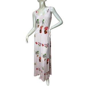 May include: A light pink sleeveless maxi dress with a floral pattern. The dress features a scoop neckline and a flowing silhouette. The floral design includes red, brown, and green elements. The dress is displayed on a mannequin.