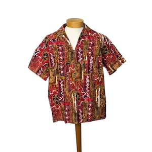 May include: Short-sleeved shirt with a vibrant geometric pattern in red, brown, black, and white. The shirt features a collared neckline and is displayed on a mannequin. The fabric has a textured appearance, suggesting a vintage or retro style.