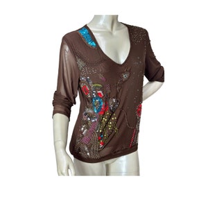Puede incluir: Top marrón de manga larga con cuello en V profundo. La tela transparente está adornada con lentejuelas, abalorios y bordados coloridos en tonos azules, rojos, verdes y dorados. El top se muestra en un maniquí.