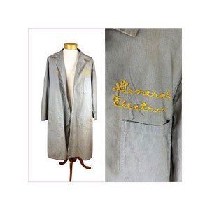 May include: A light gray vintage lab coat with the words "General Electric" embroidered in yellow thread. The coat has a pocket on the chest and is displayed on a mannequin. The coat is long-sleeved and reaches the knees.