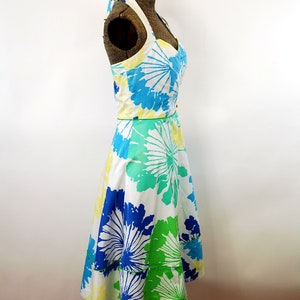 May include: A sleeveless halter dress featuring a white base with a large-scale floral print in shades of bright blue, lime green, and pale yellow. The dress has a sweetheart neckline, a fitted bodice, and a flared skirt with a horizontal seam.