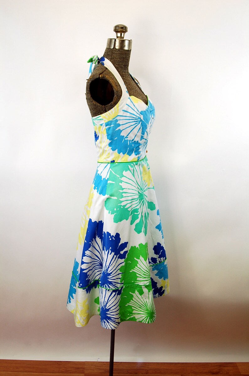 May include: A sleeveless halter dress featuring a white base with a large-scale floral print in shades of bright blue, lime green, and pale yellow. The dress has a sweetheart neckline, a fitted bodice, and a flared skirt with a horizontal seam.