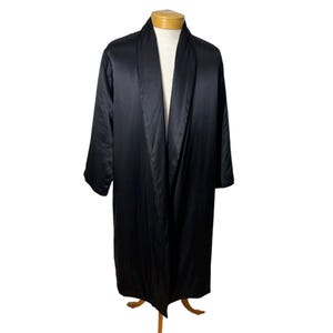 May include: A black satin robe with a shawl collar is displayed on a wooden mannequin. The robe has long sleeves and a floor-length hem. The fabric appears smooth and has a slight sheen, suggesting a luxurious feel.