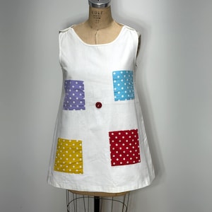 May include: A sleeveless white dress featuring four square patches with white polka dots in purple, light blue, yellow, and red. A single red button is centered on the front of the dress, which is displayed on a mannequin with a Wolf brand label.