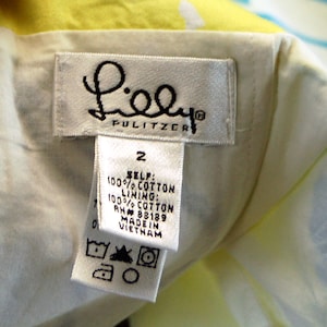 May include: A white clothing label with the text Lilly Pulitzer, size 2, 100% cotton self and lining, RN 88189, made in Vietnam, and care symbols. The label is attached to a garment with a yellow and white patterned fabric visible in the background.