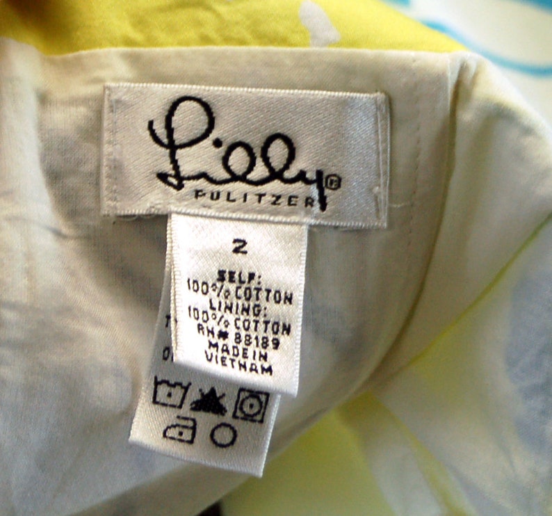May include: A white clothing label with the text Lilly Pulitzer, size 2, 100% cotton self and lining, RN 88189, made in Vietnam, and care symbols. The label is attached to a garment with a yellow and white patterned fabric visible in the background.