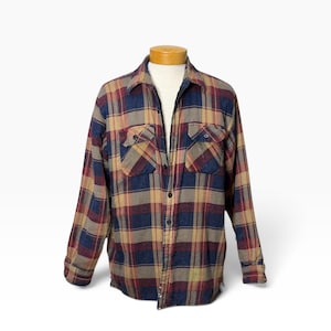 May include: A long-sleeve button-down shirt featuring a plaid pattern in shades of navy blue, burgundy, and tan. The shirt includes a standard collar, a full-length front button closure, and two chest pockets with buttoned flaps on each side.